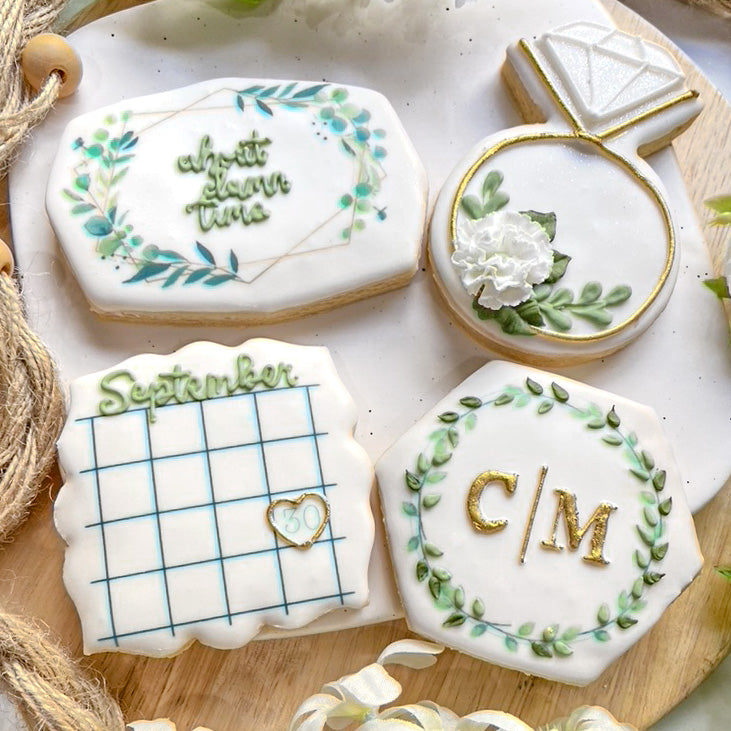 Four rectangular cookies decorated with green and white icing. The designs include the phrase "About Damn Time" surrounded by leaves, a calendar for September with a heart on the 30th, a ring with a white flower, and a hexagon with the letters "C/M" inside a leafy border.