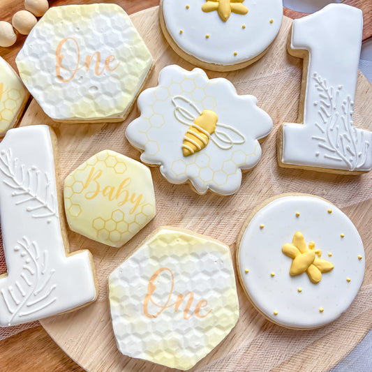 Nine sugar cookies in various shapes, including circles, hexagons, and the number one, decorated with a honey bee theme in yellow and white.