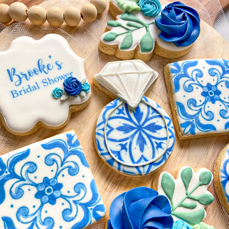 A collection of cookies decorated with blue and white patterns reminiscent of Portuguese tiles. The cookies include a ring, floral accents, and a plaque that reads "Brooke's Bridal Shower."