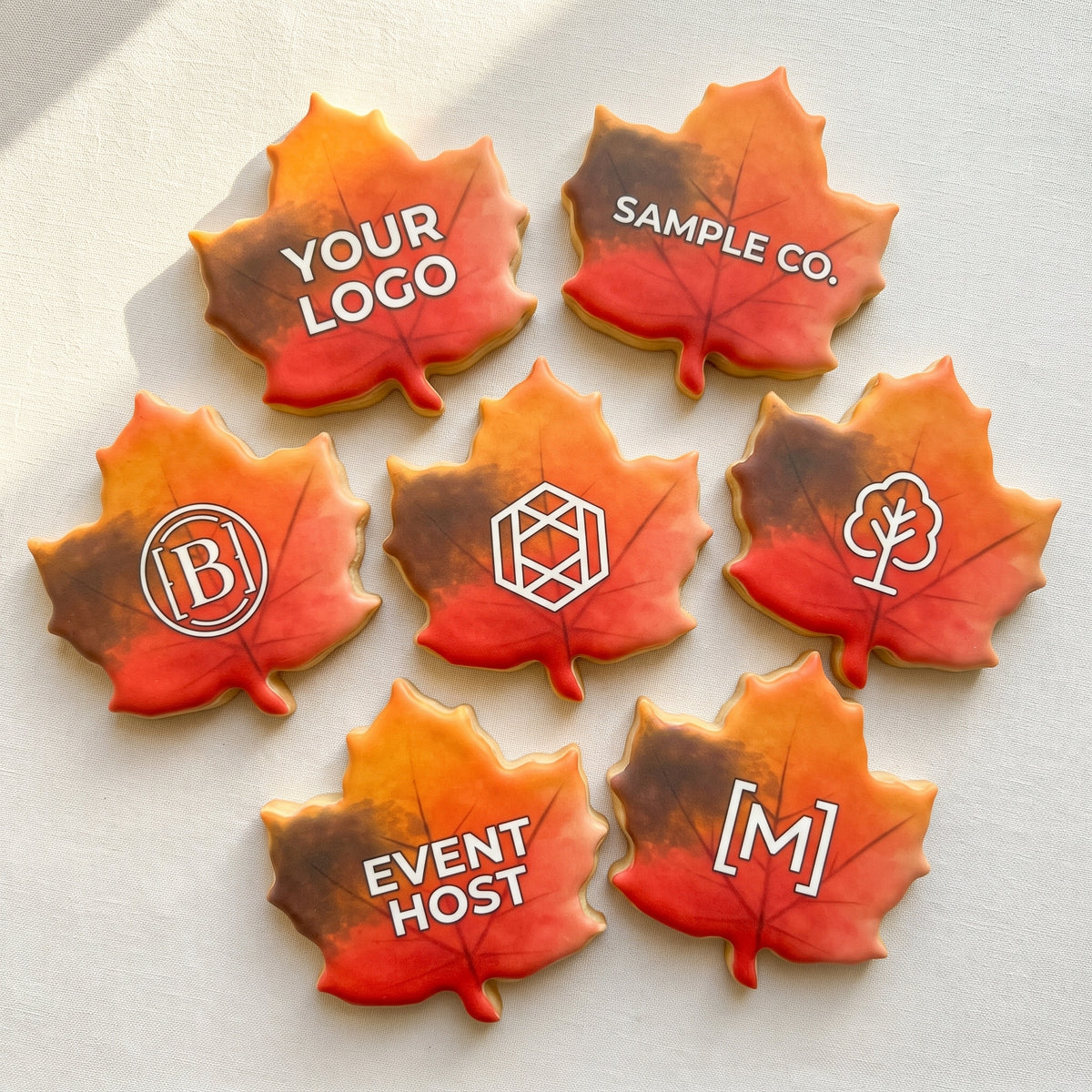 A maple leaf shaped sugar cookie with a hand-painted red-to-orange-to-gold autumn ombre and a custom printed company logo centered on top.