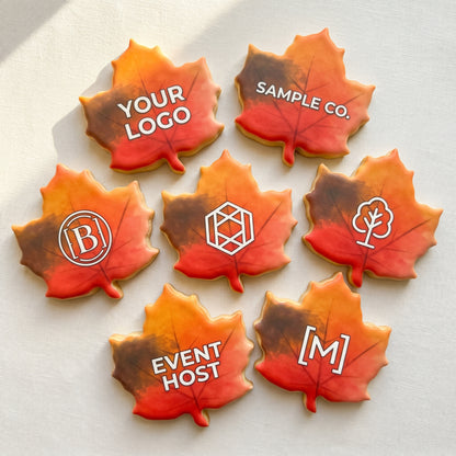 A maple leaf shaped sugar cookie with a hand-painted red-to-orange-to-gold autumn ombre and a custom printed company logo centered on top.
