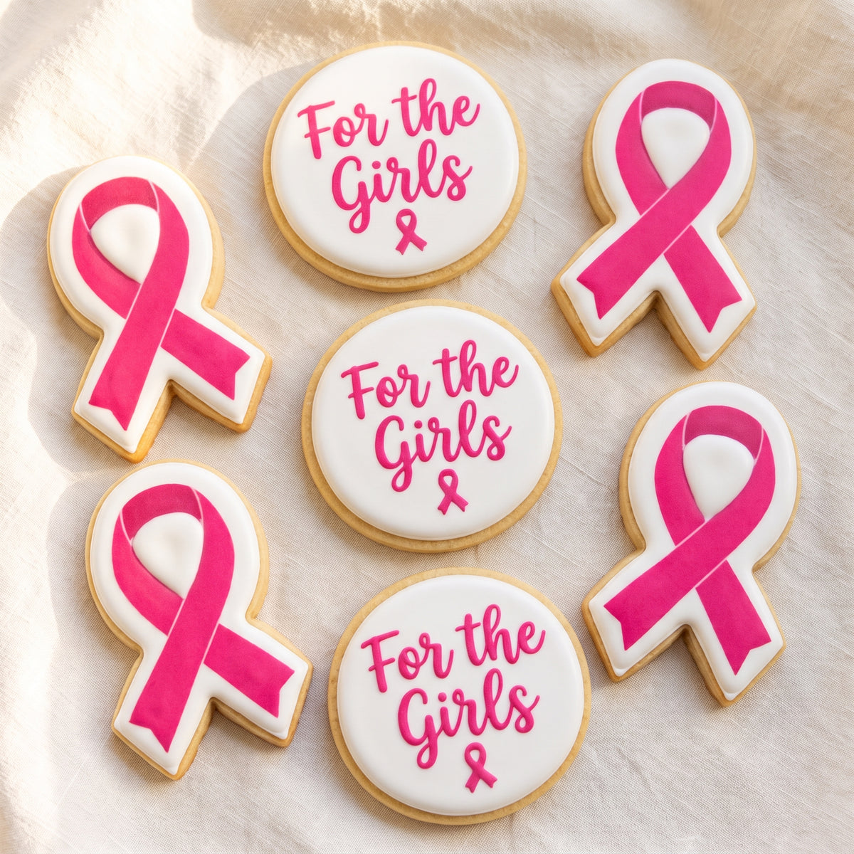 A flat lay of six awareness cookies — three ribbon-shaped cookies with pink ribbons printed on white and three white circles with hot pink 'For the Girls' script and small pink ribbon details.
