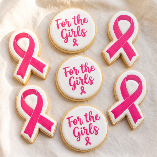 A flat lay of six awareness cookies — three ribbon-shaped cookies with pink ribbons printed on white and three white circles with hot pink 'For the Girls' script and small pink ribbon details.